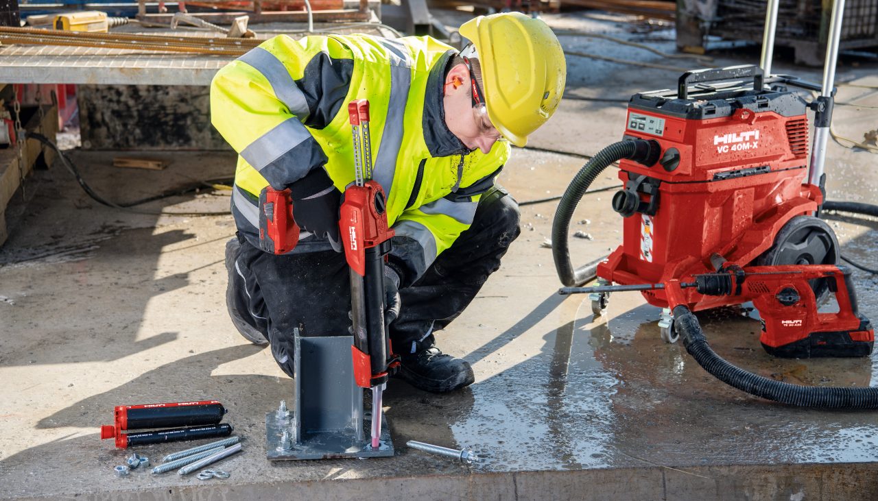 Construction worker in yellow high visibility vest injecting chemical anchor into a drill hole in concrete.
